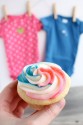 A woman holding a cupcake above a couple baby outfits.